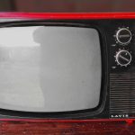 A vintage television. The edges of the set are red, and the screen is off. There are dials on the top right of the set. Christmas TV